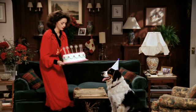 A woman in red carrying a birthday cake beside a dog in a party hat