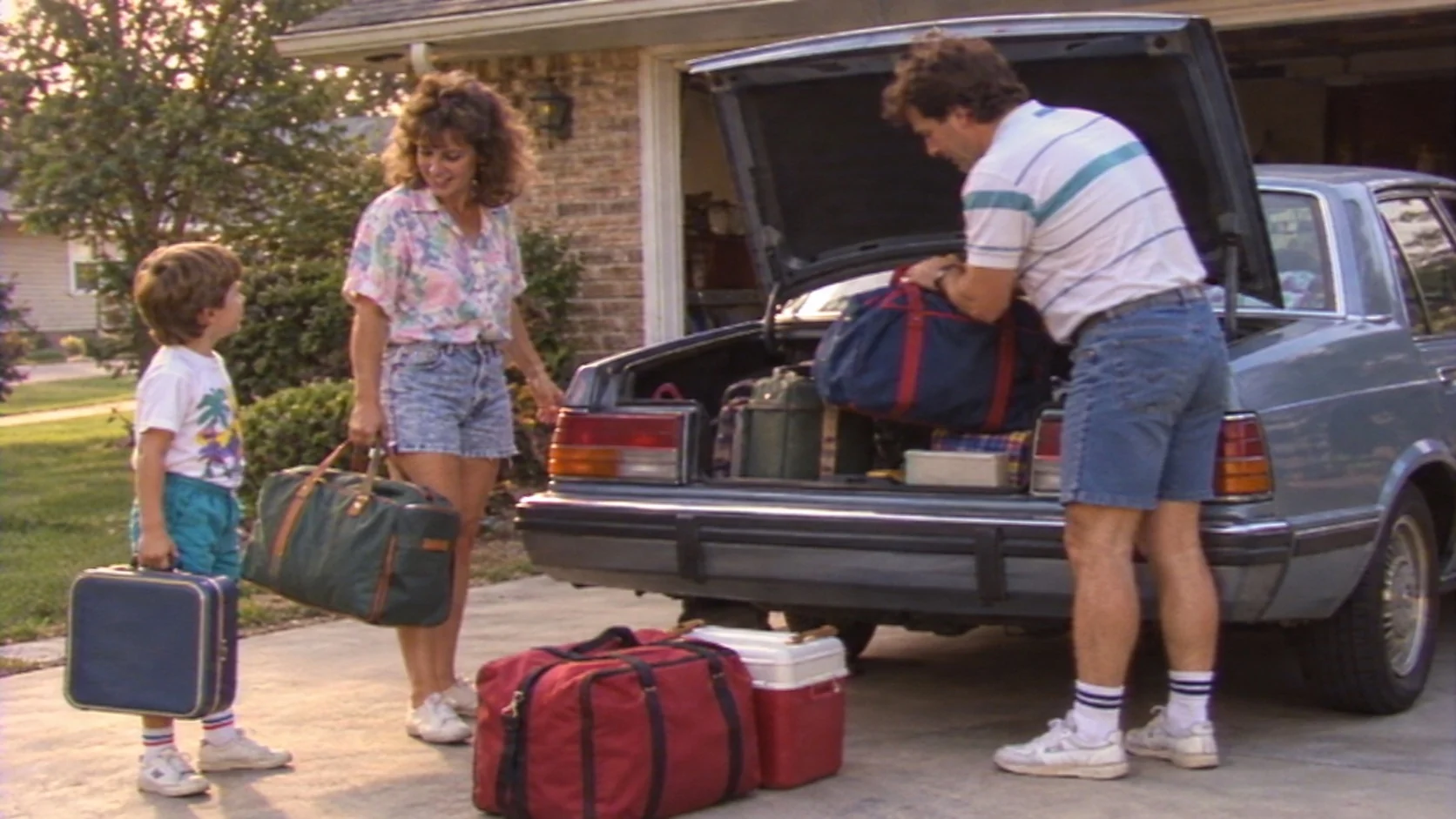 Late-1980s family packs suitcases into a car for a summer trip, shown with a soft handheld VHS home-video look